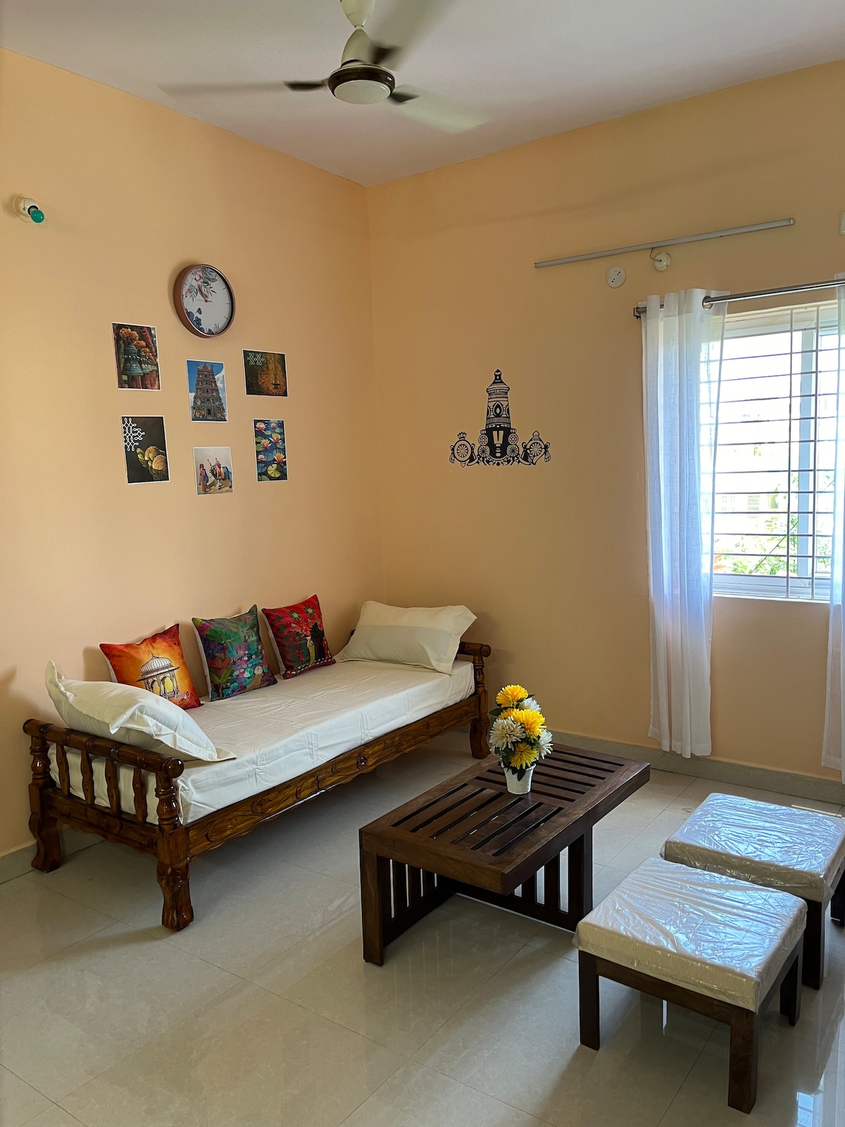 A bright sitting area is featured, with a wooden daybed adorned with colorful cushions. A small wooden coffee table is positioned in front, accompanied by two light-colored stools. On the wall, an assortment of framed artworks and a clock enhance the inviting ambiance, while natural light filters through a window.