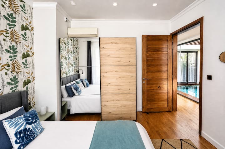Ambiance cosy de la chambre 2 : chevet bois, lampe de lecture et lit 180x200 avec coussins déco. Papier peint tropical exclusif. Grand dressing