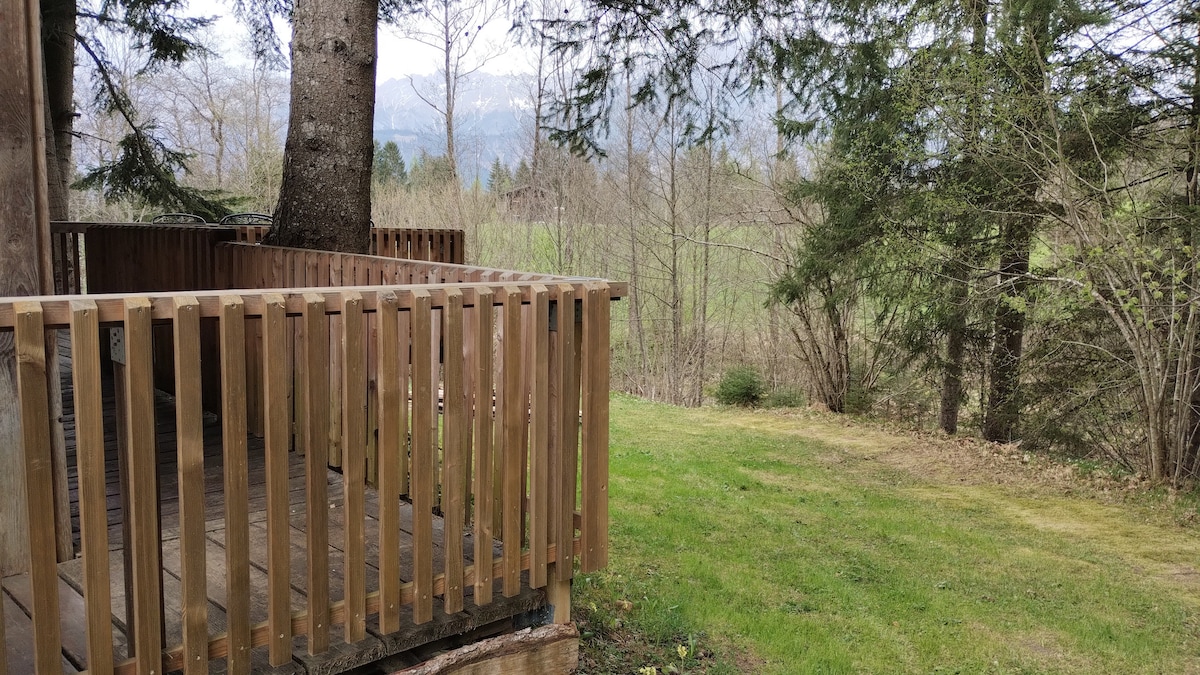 A wooden terrace features a railing made of vertical slats, offering views of the surrounding greenery and distant mountainous landscape. The area is surrounded by trees, with a grassy patch visible nearby.