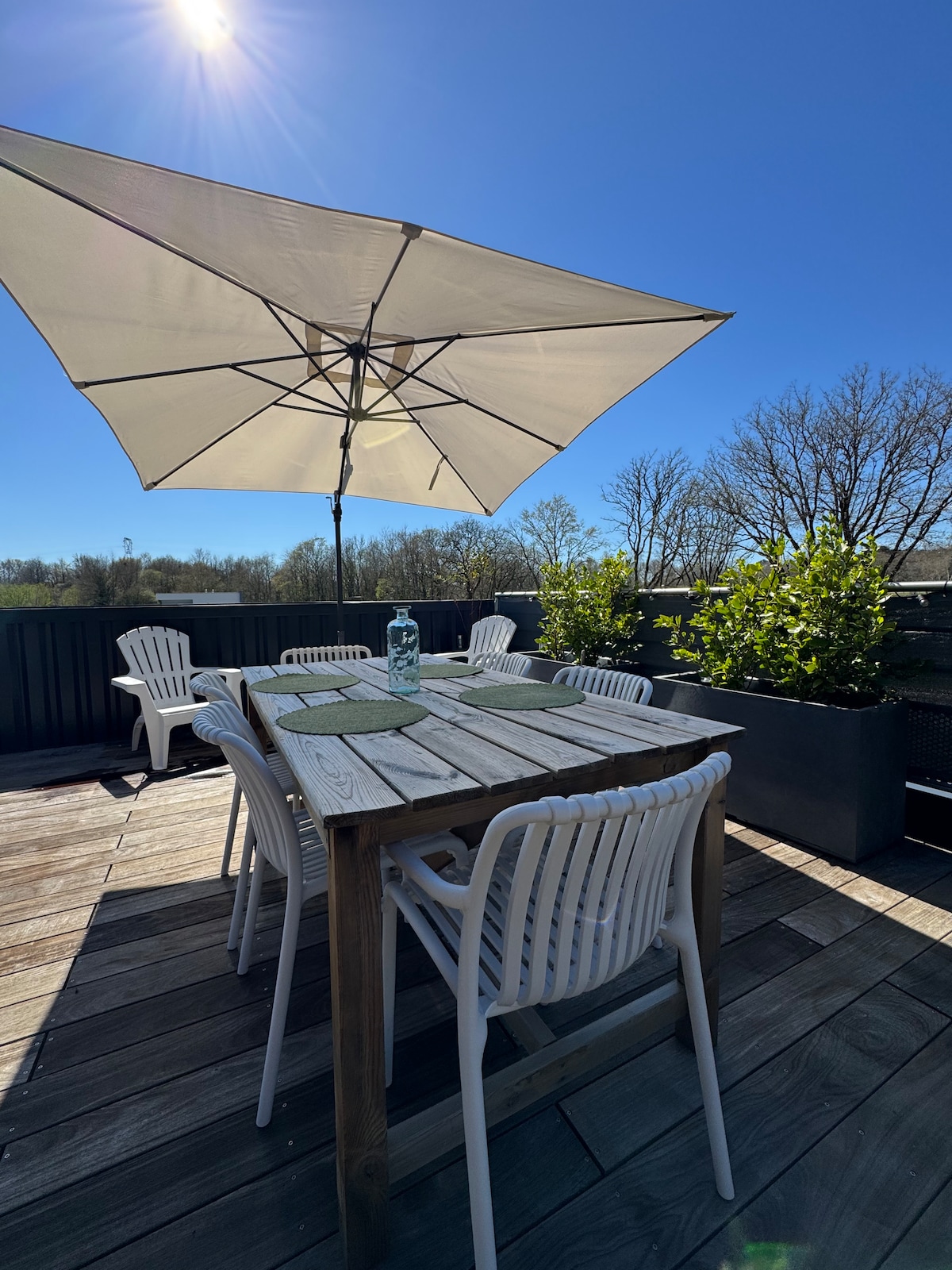 A spacious terrace is displayed under a bright blue sky, featuring a wooden dining table surrounded by several white chairs. An umbrella provides shade, and potted plants are placed around the perimeter, enhancing the outdoor setting.