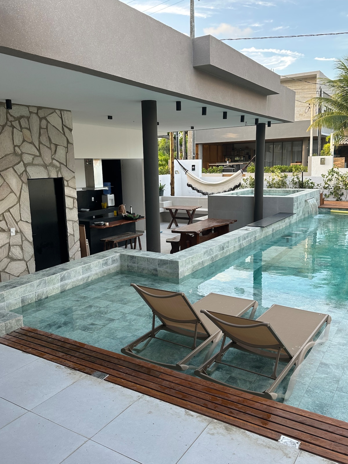An inviting outdoor space features a turquoise pool adjacent to a lounge area with two sun loungers. A well-equipped kitchen and dining area are visible in the background, while a hammock adds a relaxing touch. Lush greenery surrounds the area, enhancing the tranquil setting.