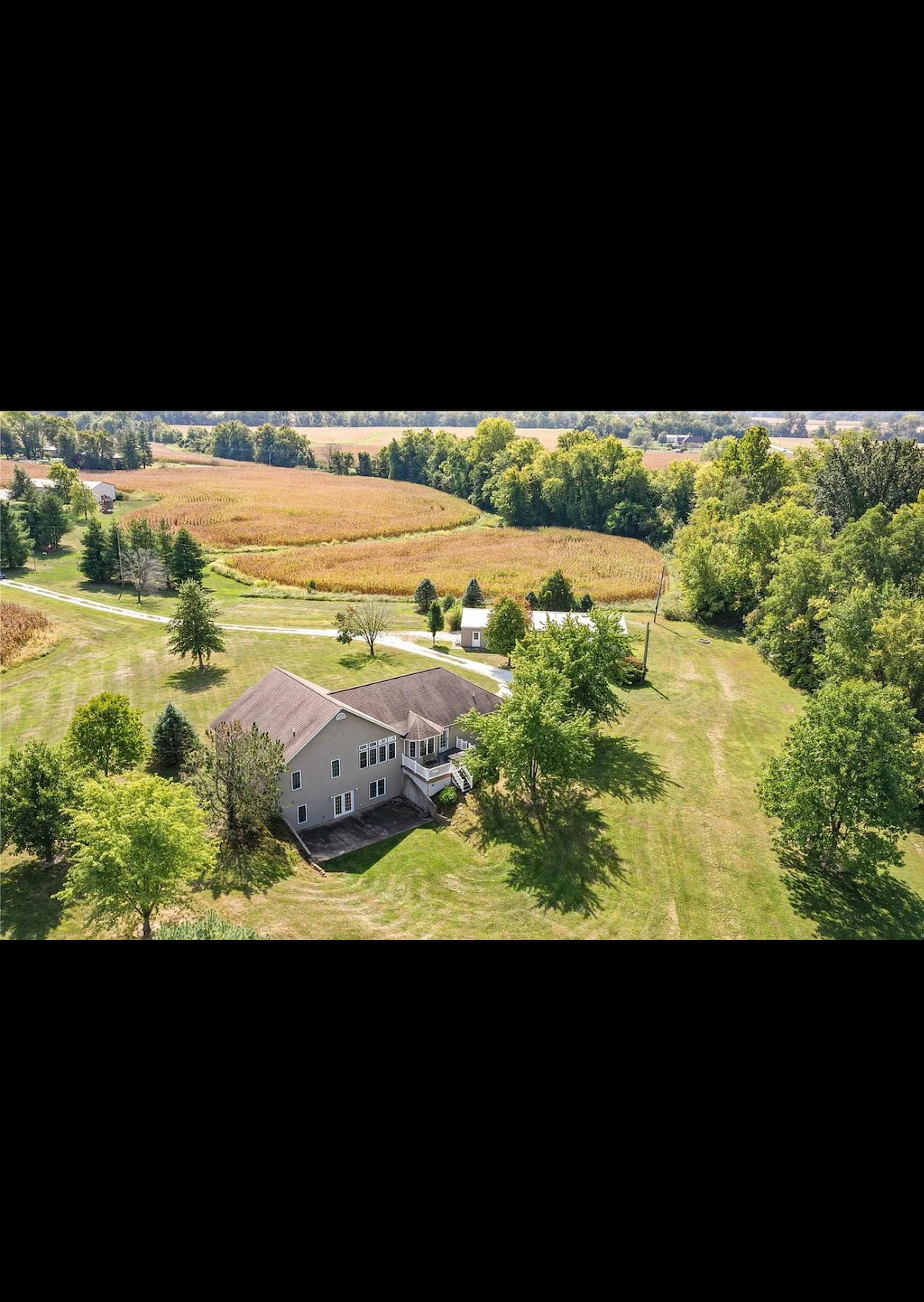 An aerial view captures the large Countryside property, surrounded by expansive greenery and fields. The two-story guesthouse is visible, with a well-maintained yard featuring trees and an inviting pathway leading to nearby facilities. The peaceful rural landscape enhances the serene setting.