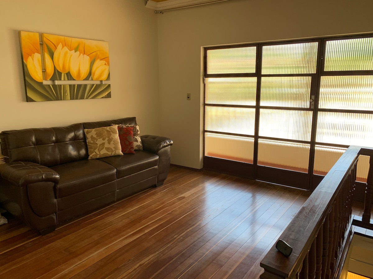 A spacious living area is furnished with a brown leather sofa adorned with decorative pillows. Large windows featuring textured glass panels allow natural light to fill the room, casting soft reflections onto the polished wooden floor.
