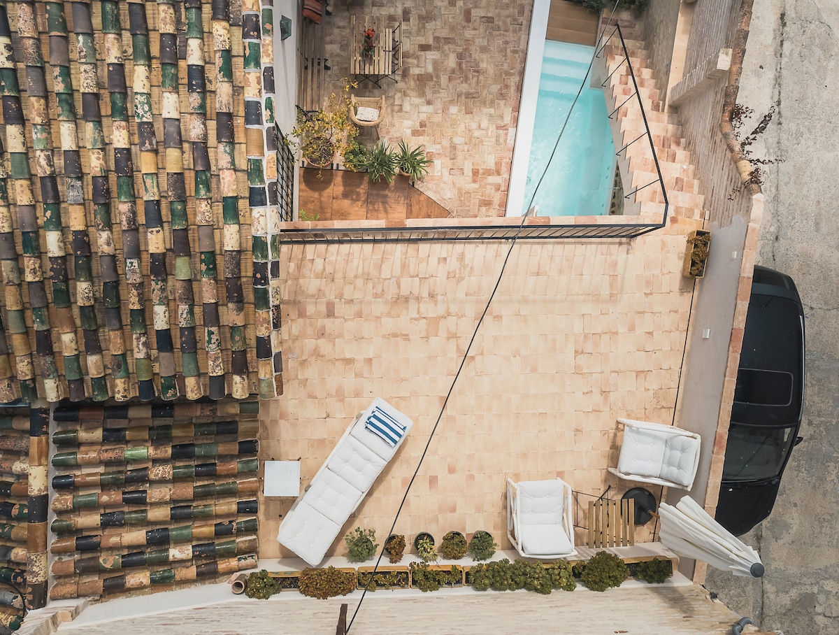 An aerial view displays a serene outdoor area featuring a pool bordered by terracotta tiles. Comfortable seating arrangements, including two white loungers and a shaded area, create a relaxing environment. Lush greenery and decorative plants are strategically placed to enhance the space.