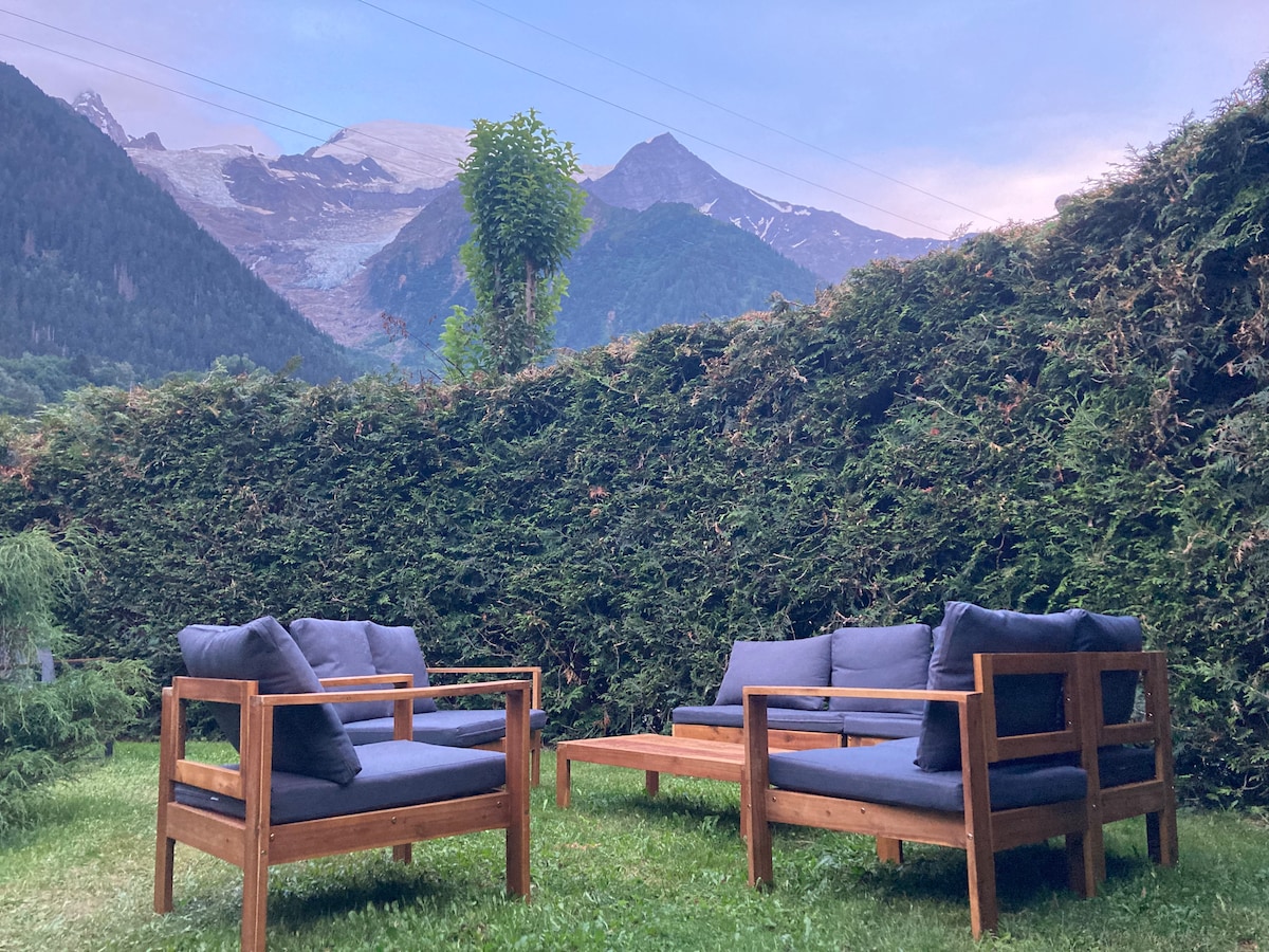A seating arrangement featuring two wooden chairs and a coffee table sets on a grassy area. Green hedges provide privacy, while a backdrop of mountains and a glacier is visible in the distance, contributing to a serene outdoor setting.