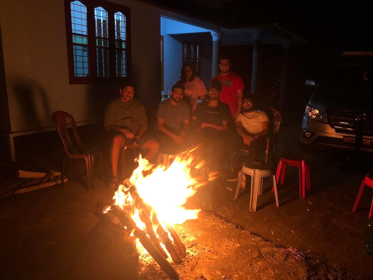 A campfire is actively burning in front of seated individuals, providing warmth and light in the evening. Surrounding chairs are arranged in a semi-circle, with soft shadows created by the fire's glow. The background features a structure with windows illuminated by soft light.