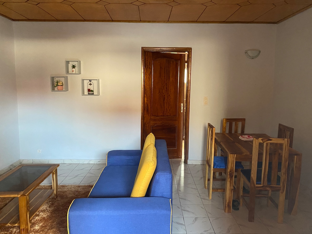An open living area is presented, featuring a blue sofa with yellow cushions. A wooden dining table with matching chairs is situated adjacent to the sofa. Light-colored tiled flooring adds to the spacious feel, while decorative frames are displayed on the wall.