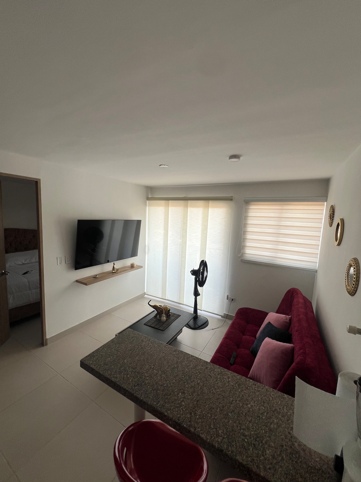 A bright living area is shown, featuring a red sofa, flat-screen television mounted on the wall, and a small table with decorative elements. Natural light enters through sheer window coverings, and a fan is positioned nearby, offering comfort in the space.