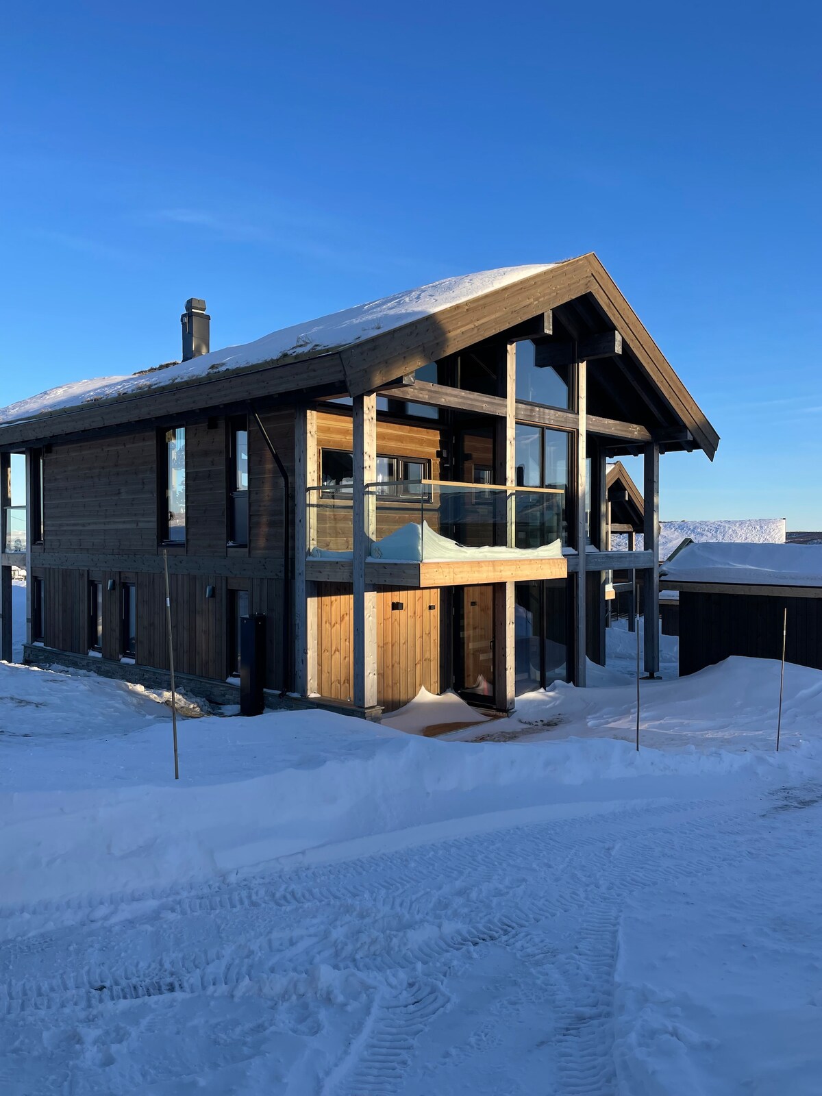 A modern two-story cabin is displayed, constructed with natural wood materials. Large glass doors and windows are visible, allowing ample sunlight to illuminate the interior. Snow blankets the ground, creating a serene winter setting, with a clear blue sky above.