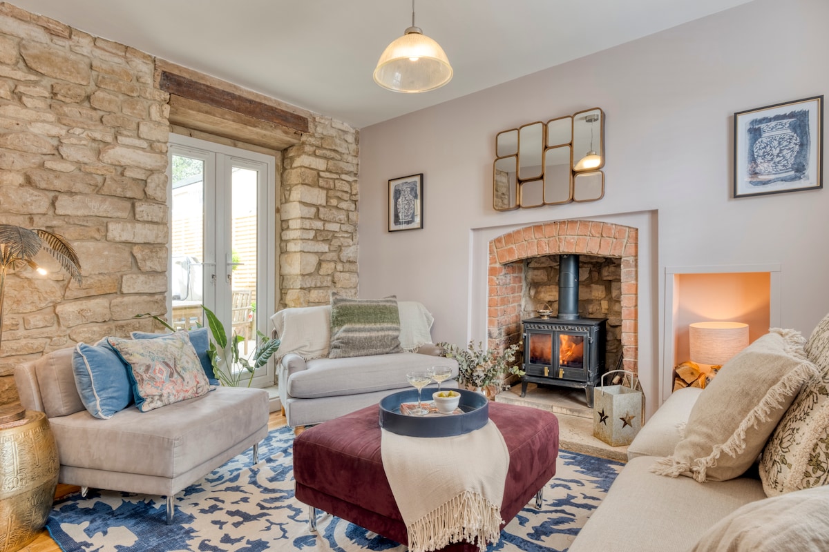 A warm living area features exposed stone walls and a cozy fireplace with a wood-burning stove. Light-colored sofas are arranged around a decorative coffee table, with a soft rug beneath. Subtle artwork decorates the walls, and natural light filters in through the doors.