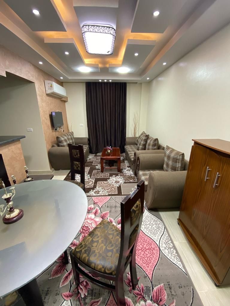 A comfortable seating area is highlighted by a patterned rug. Two armchairs and a sofa provide ample seating, complemented by a small wooden table at the center. A dining table with decorative chairs is positioned near the wall, and soft lighting enhances the inviting ambiance.