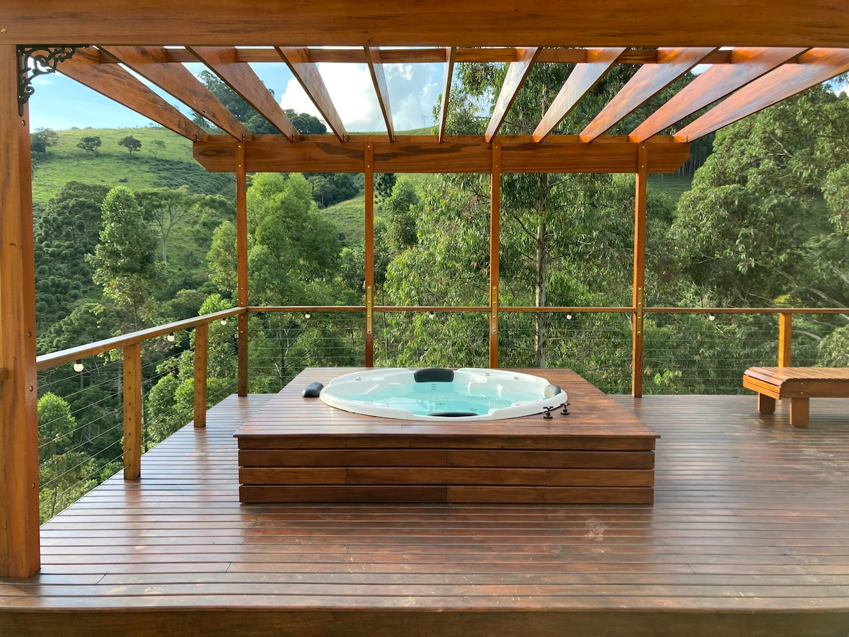 A spacious wooden deck is surrounded by lush greenery, featuring a hot tub positioned on a wooden platform. The open design allows for unobstructed views of the mountain landscape, creating a tranquil outdoor space for relaxation.