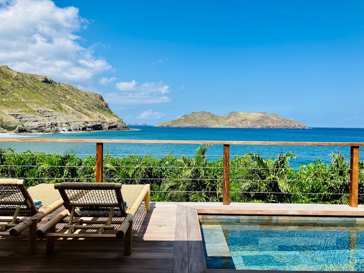 A serene terrace features a private pool overlooking the Atlantic Ocean. Two lounge chairs are positioned towards the water, providing views of distant islands against a bright blue sky. Lush greenery surrounds the area, enhancing the tranquil setting.