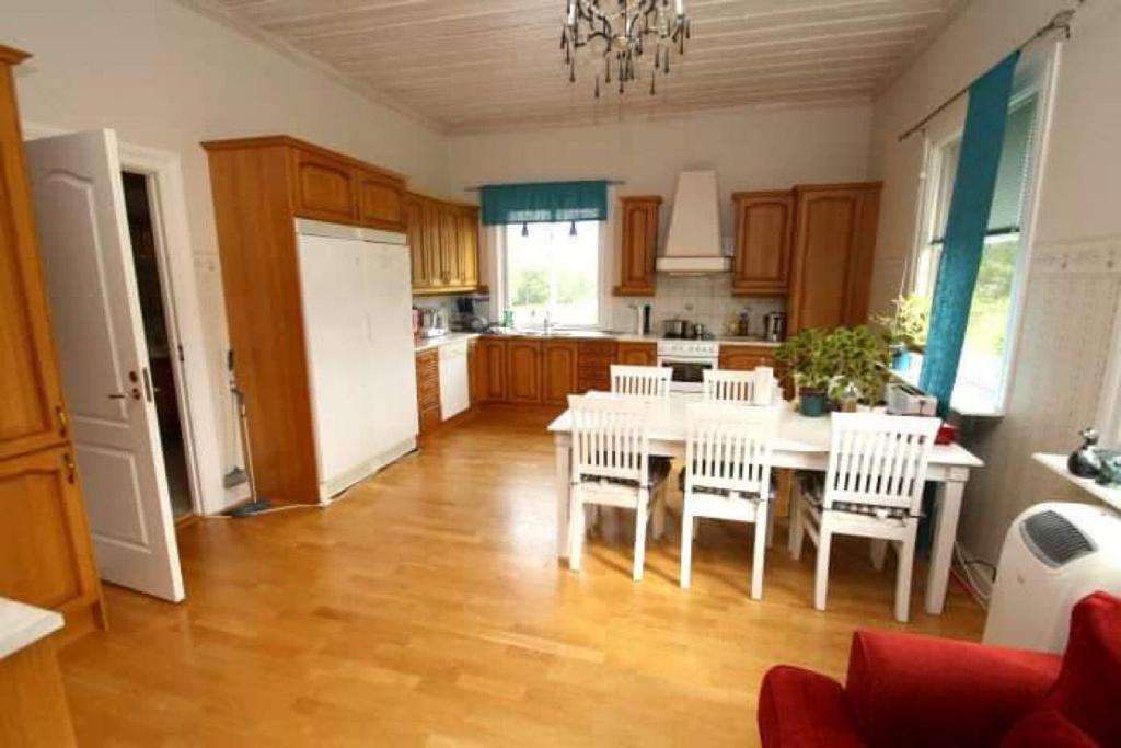 A spacious kitchen is featured, showcasing wooden cabinetry and a large white dining table surrounded by chairs. Natural light enters through a window, highlighting the warm wooden flooring. A small plant sits on the countertop, adding a touch of greenery to the neutral color scheme.