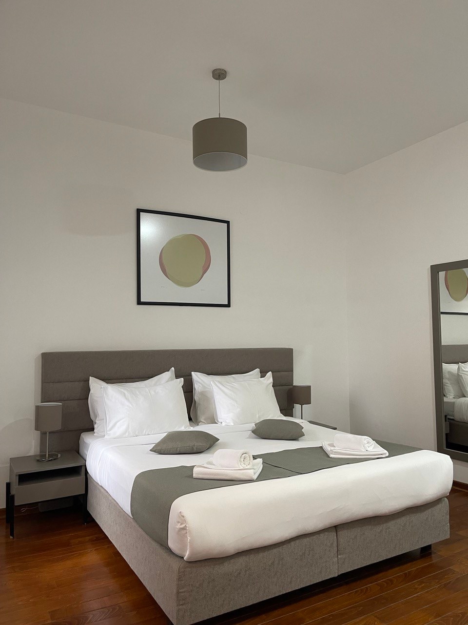A spacious bedroom is furnished with a large bed containing soft white linens and gray accent pillows. Adjacent nightstands support neatly arranged lamps. A mirror reflects the room's simplicity, while artwork hangs above the bed, contributing to the serene aesthetic.