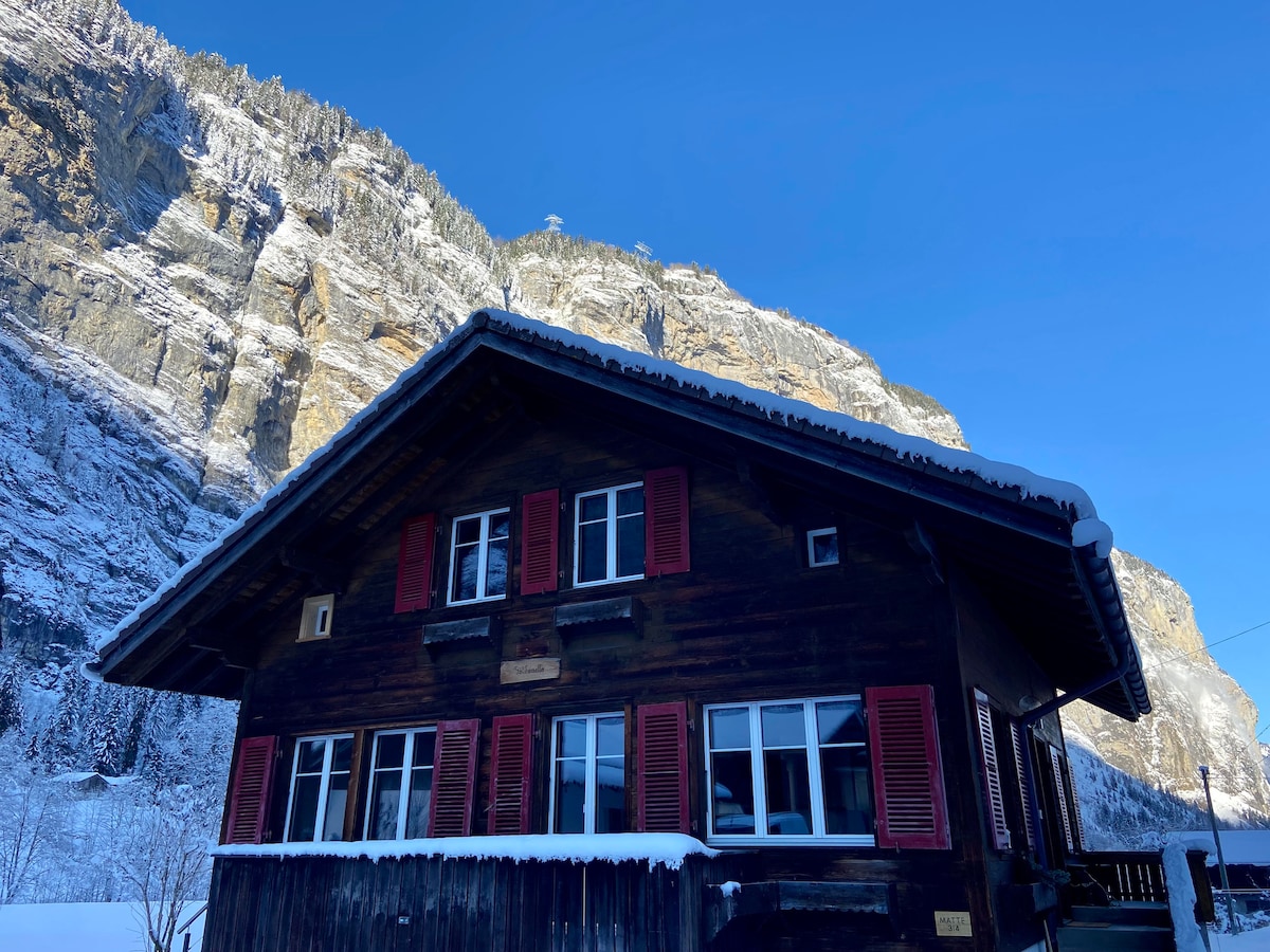 A traditional Swiss chalet features dark wooden walls and bright red shutters, set against a backdrop of snow-covered mountains. The structure is adorned with multiple windows, allowing for ample natural light, while a clear blue sky complements the serene winter landscape.
