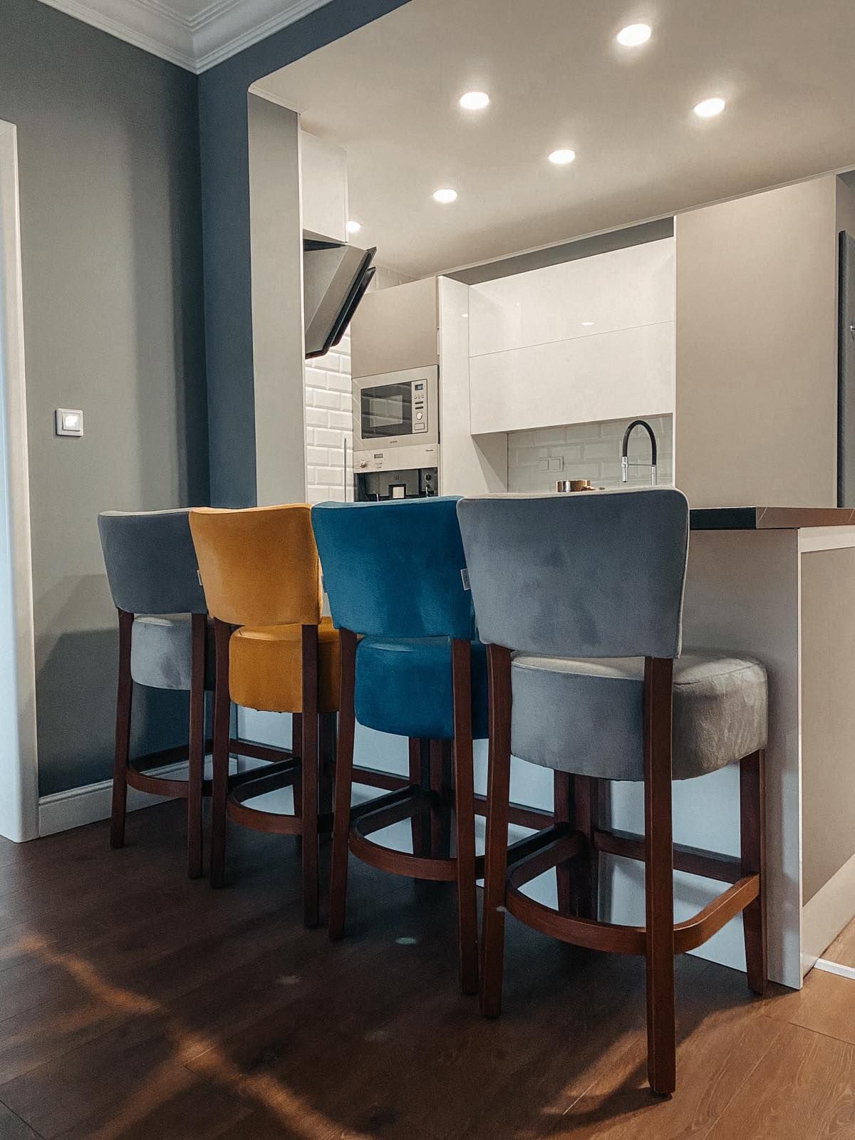 A sleek kitchen bar area features four upholstered stools in varying colors, including gray, mustard yellow, and teal. The countertops and cabinetry exhibit a modern design, while overhead lighting creates a well-lit space, contributing to the functional kitchen atmosphere.