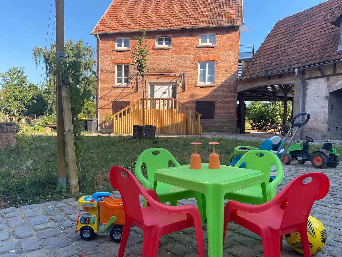 A playful outdoor area is presented, featuring a brightly colored green table surrounded by four red chairs. Nearby, children's toys such as a colorful truck and sports balls are visible, set against the backdrop of a charming stone house with a rustic façade.