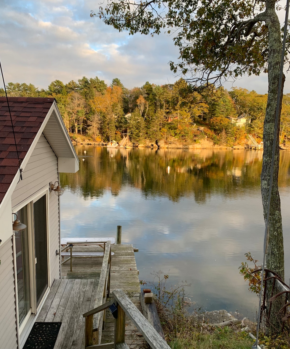A wooden dock extends from the cottage to the calm waters of Lobster Cove, reflecting the surrounding trees and autumn foliage. The image captures the tranquil ambiance, with soft lighting highlighting the peaceful setting, perfect for enjoying nature.