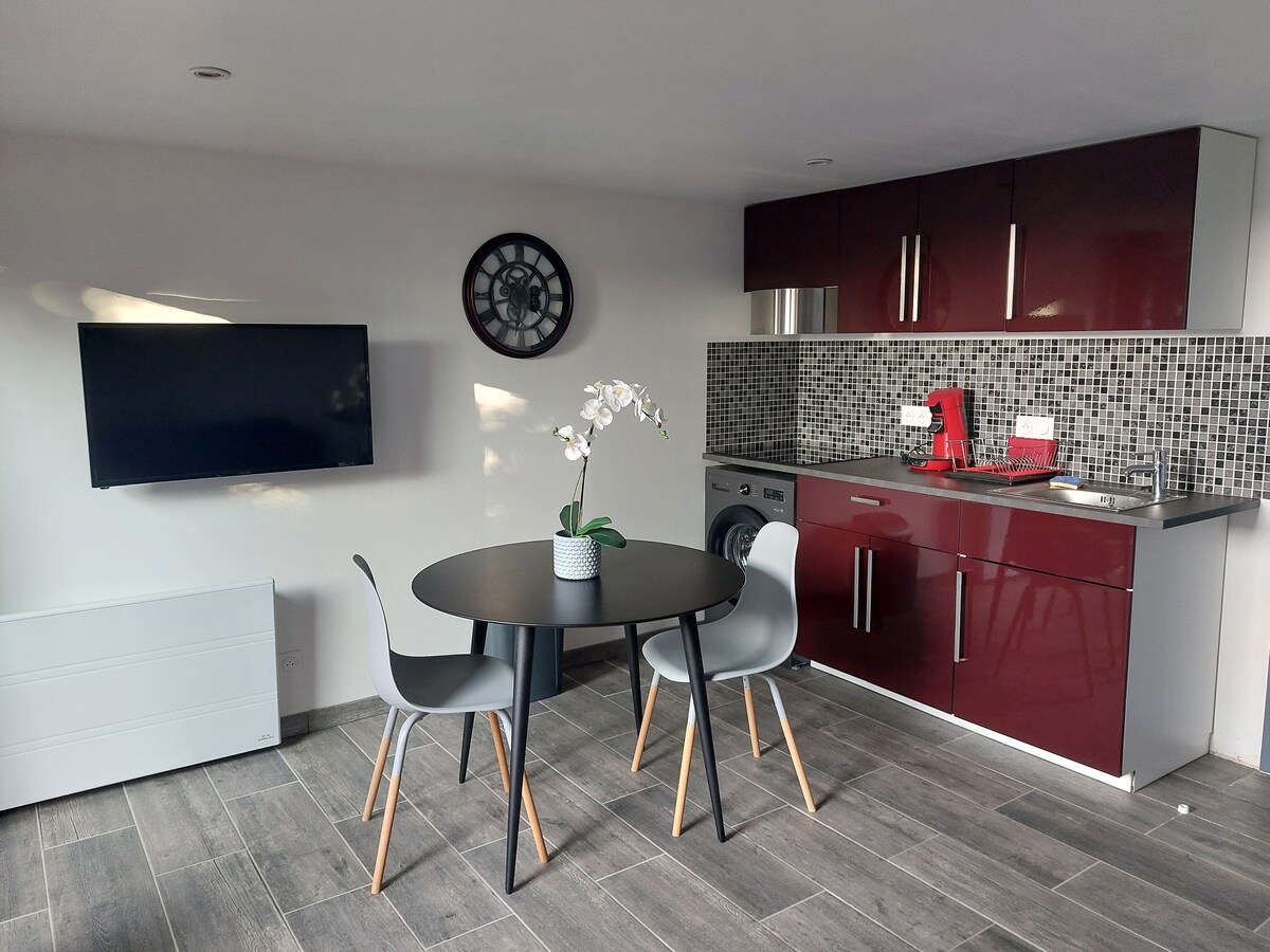 A modern kitchen area is highlighted by sleek red cabinetry and a mosaic backsplash. A round dining table with two chairs is positioned nearby. A wall-mounted television adds convenience to the space. Natural light enhances the ambiance through the large window.