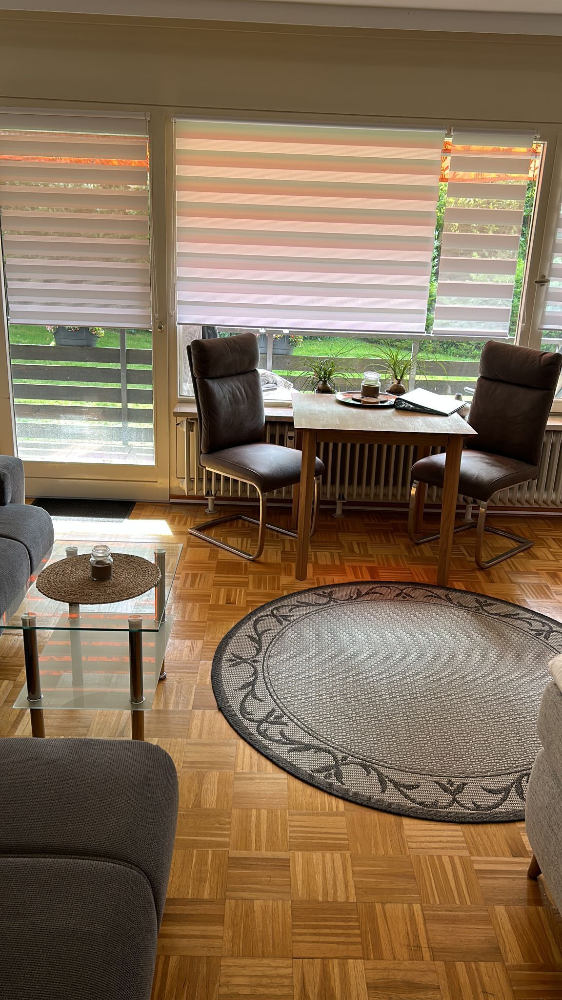 A comfortable seating area features two chairs paired with a small table, situated beside large windows adorned with striped blinds. Natural light filters through, illuminating the warm wood flooring and a circular area rug that adds texture to the space.