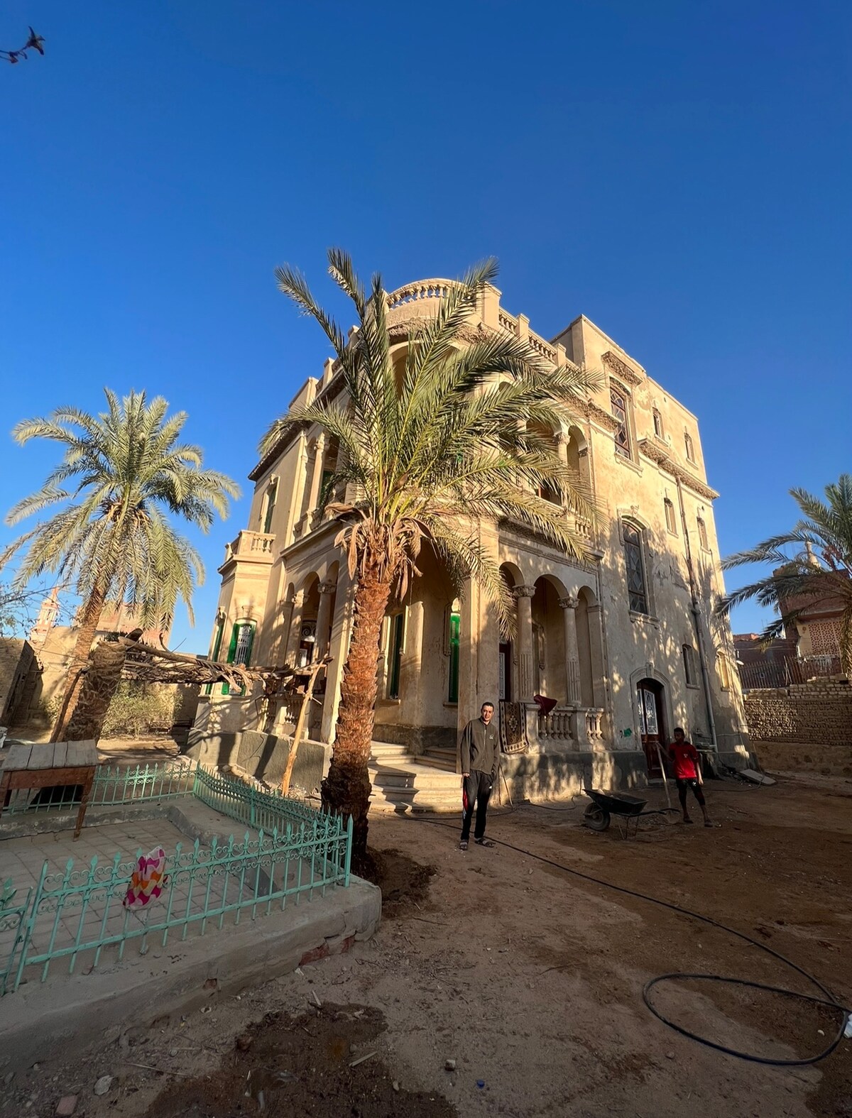 A historic building is surrounded by palm trees, showcasing a grand facade with multiple arched windows and balconies. The exterior features intricate architectural details, while a garden area lies in the foreground, with a gravel path leading towards the entrance.