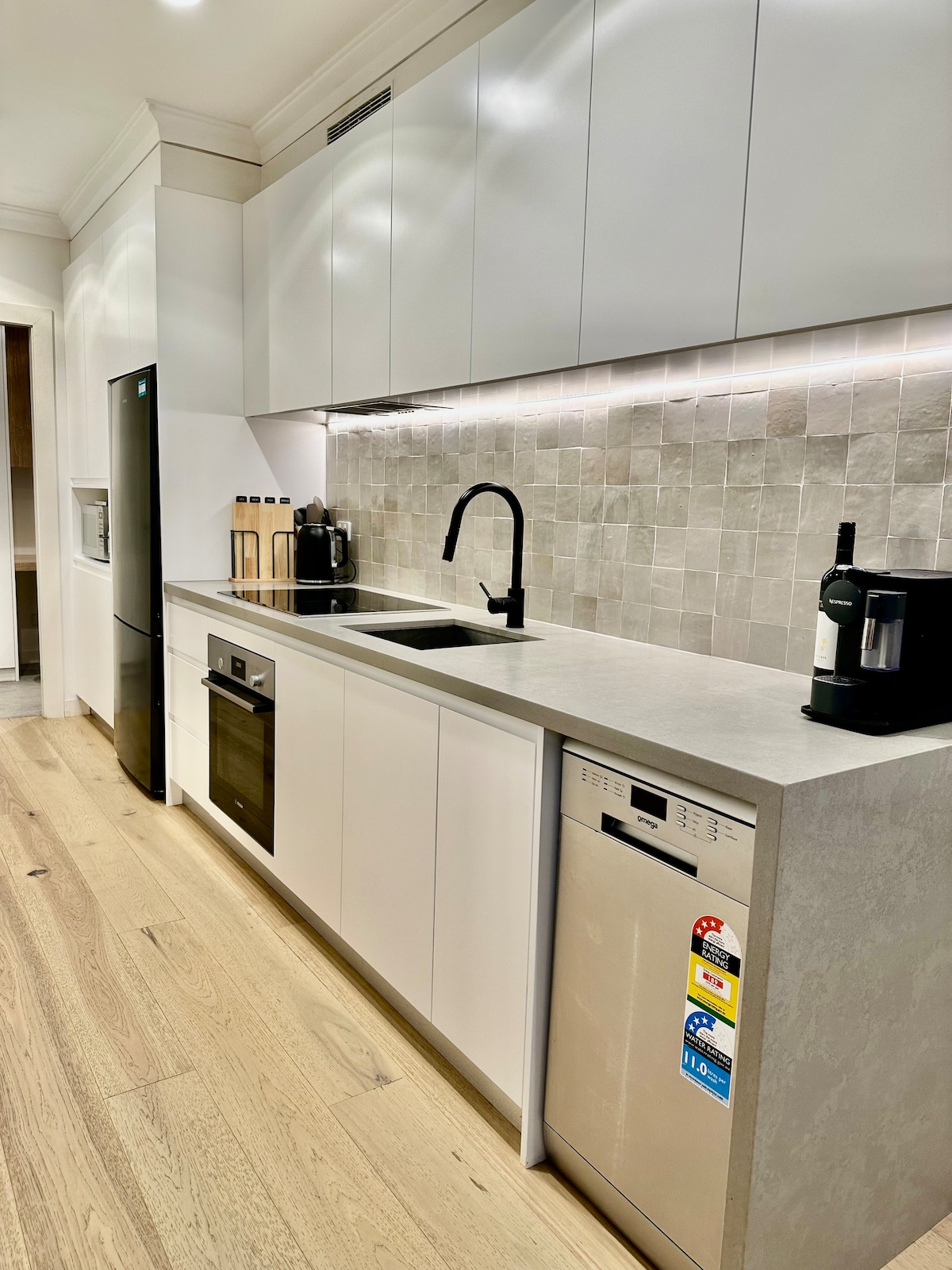 A modern kitchen is featured, showcasing sleek white cabinetry and a stone countertop. State-of-the-art appliances are included, such as an oven, microwave, and dishwasher. A stylish black faucet is positioned above the slim sink, with a coffee machine visible on the counter.