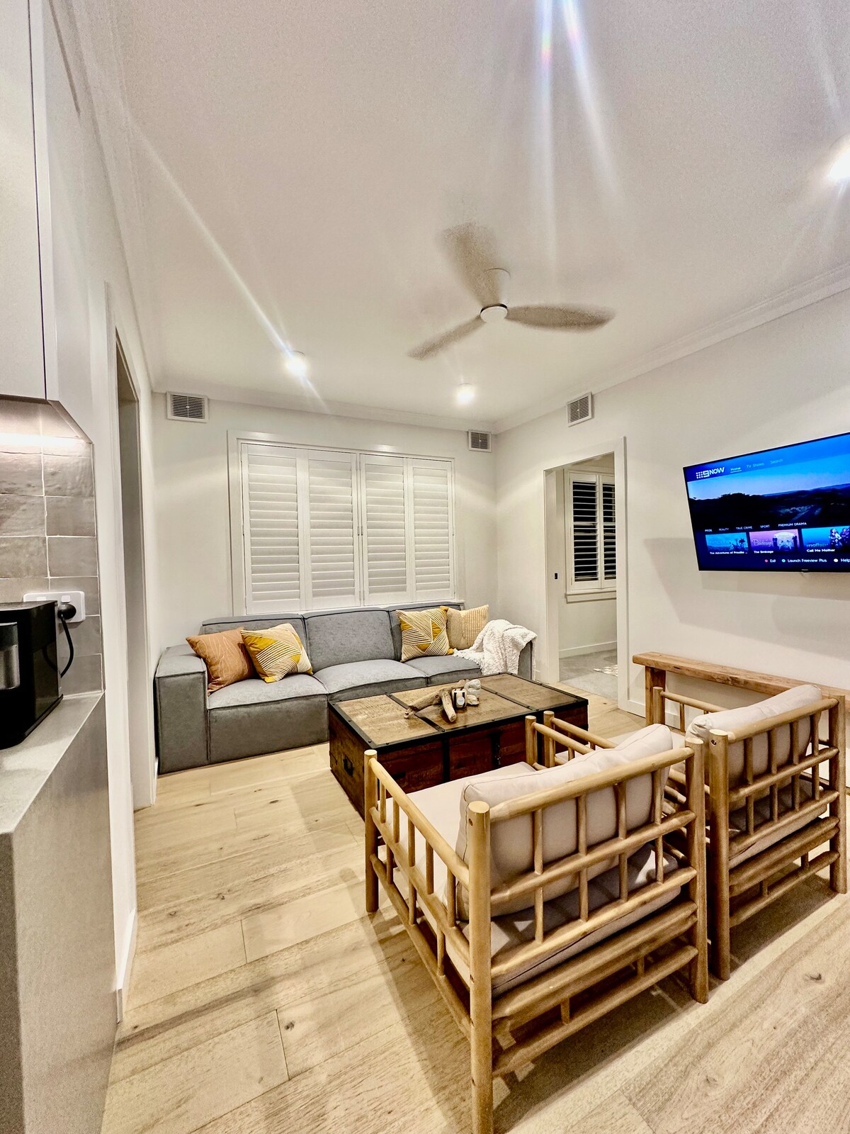 A modern living area is depicted, featuring a comfortable gray sofa with throw pillows and two bamboo chairs positioned around a wooden coffee table. A wall-mounted TV is visible, and natural light enters through the window, softly illuminating the space.