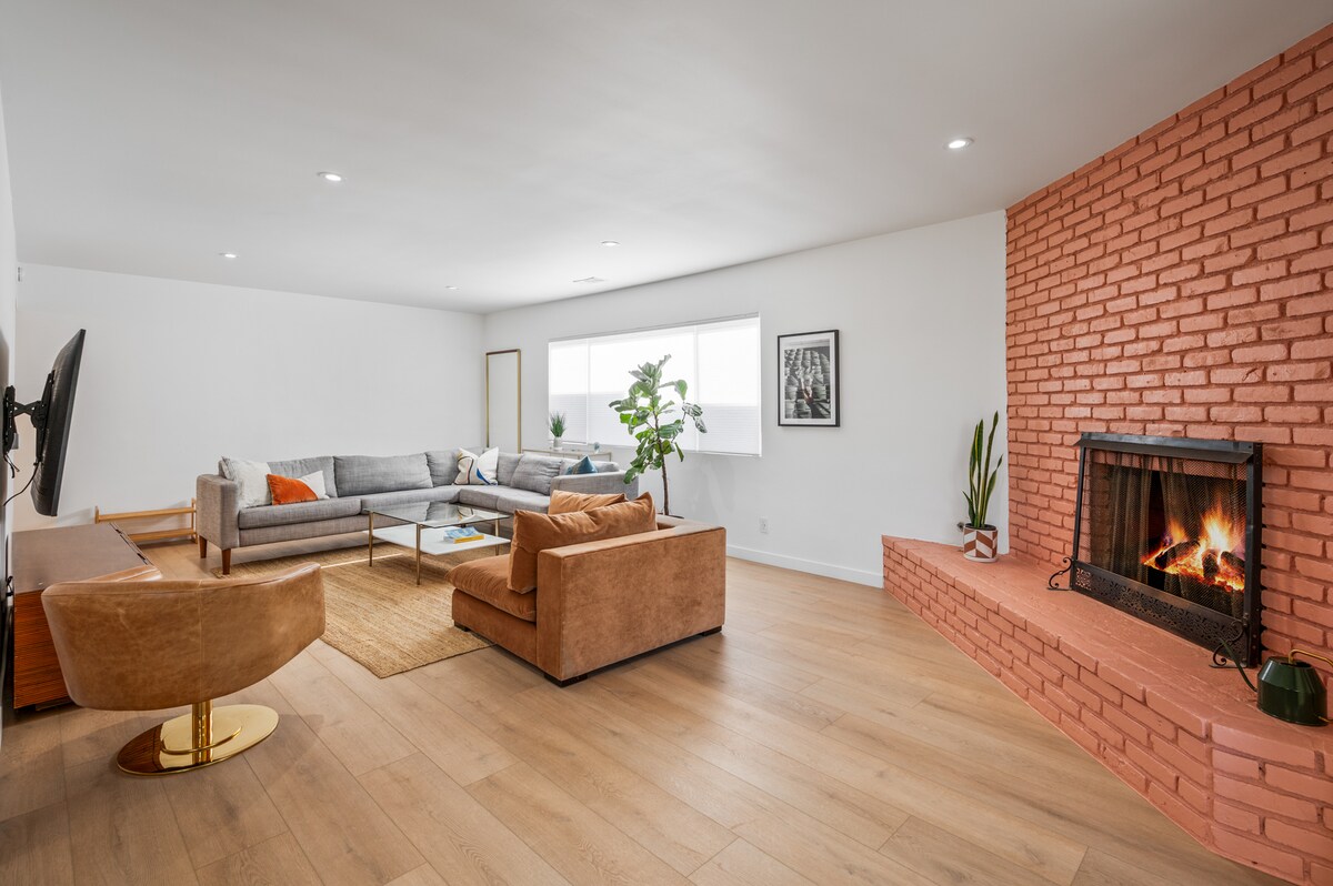 A spacious living area features a comfortable arrangement of seating, including a grey sectional and two brown chairs. A modern coffee table sits centrally, and a warm brick fireplace is showcased along one wall. Natural light fills the room through large windows.