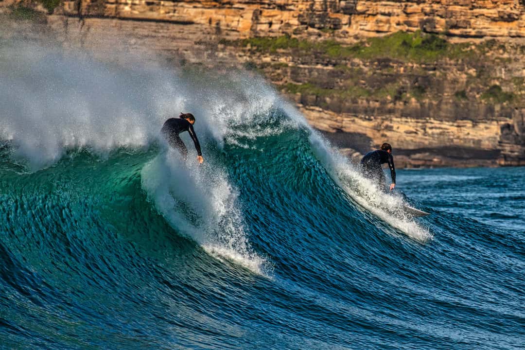 Two surfers navigate the waves in a vibrant blue ocean, with the sun catching the spray from the water. The backdrop features rugged cliffs, showcasing natural textures and colors of the coastal landscape.