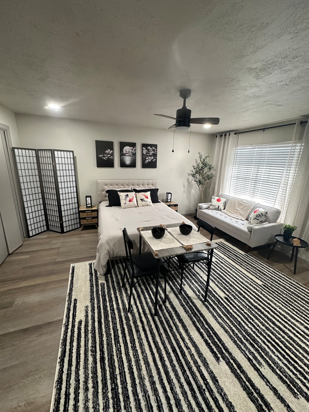 A spacious room is showcased with a large bed adorned with decorative pillows. A cozy sitting area includes a loveseat, complemented by a small dining table. Natural light filters through blinds on a large window, while decorative screens add privacy to the inviting space.