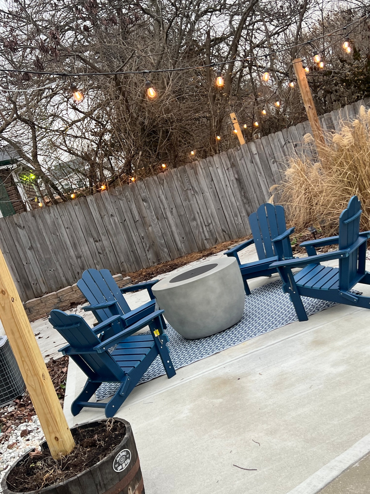 A cozy outdoor lounge area is featured, with four blue Adirondack chairs arranged around a circular fire pit. String lights hang above, adding warmth to the space. The ground is paved, and a wooden fence provides privacy, framed by tall grasses in planters.