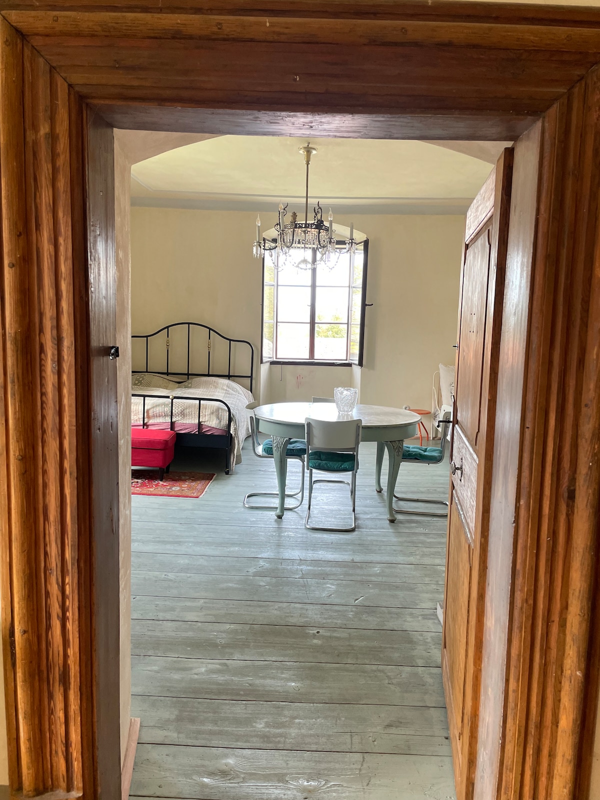 A doorway frames a spacious room featuring a cozy sleeping area with a wrought iron bed and an inviting chandelier. A circular dining table is accompanied by chairs, and sunlight filters through a large window, enhancing the overall brightness of the space.