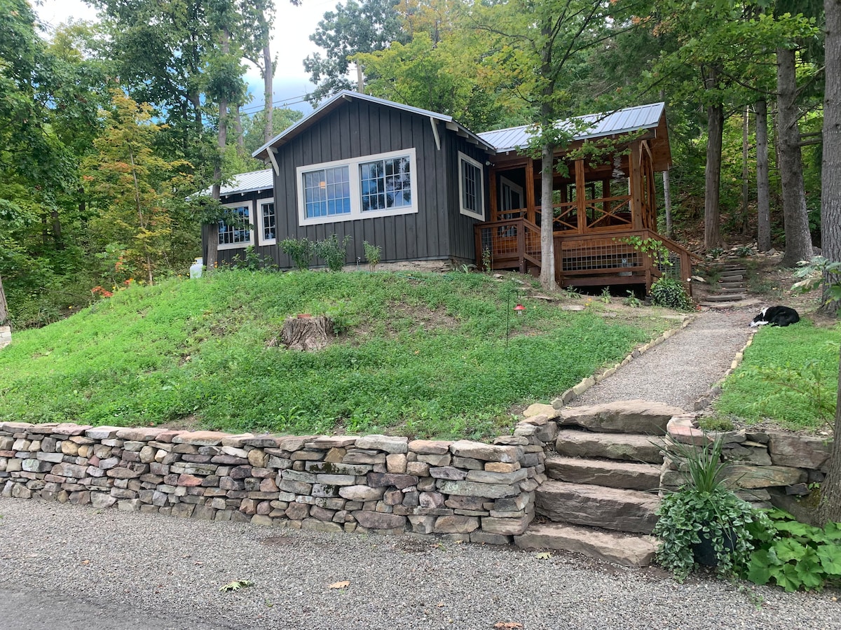 The cottage is nestled on a hillside and surrounded by trees, featuring a dark exterior with white-framed windows. A stone wall lines the front, and a pathway leads to a set of steps. The screened porch is accessible from the pathway.