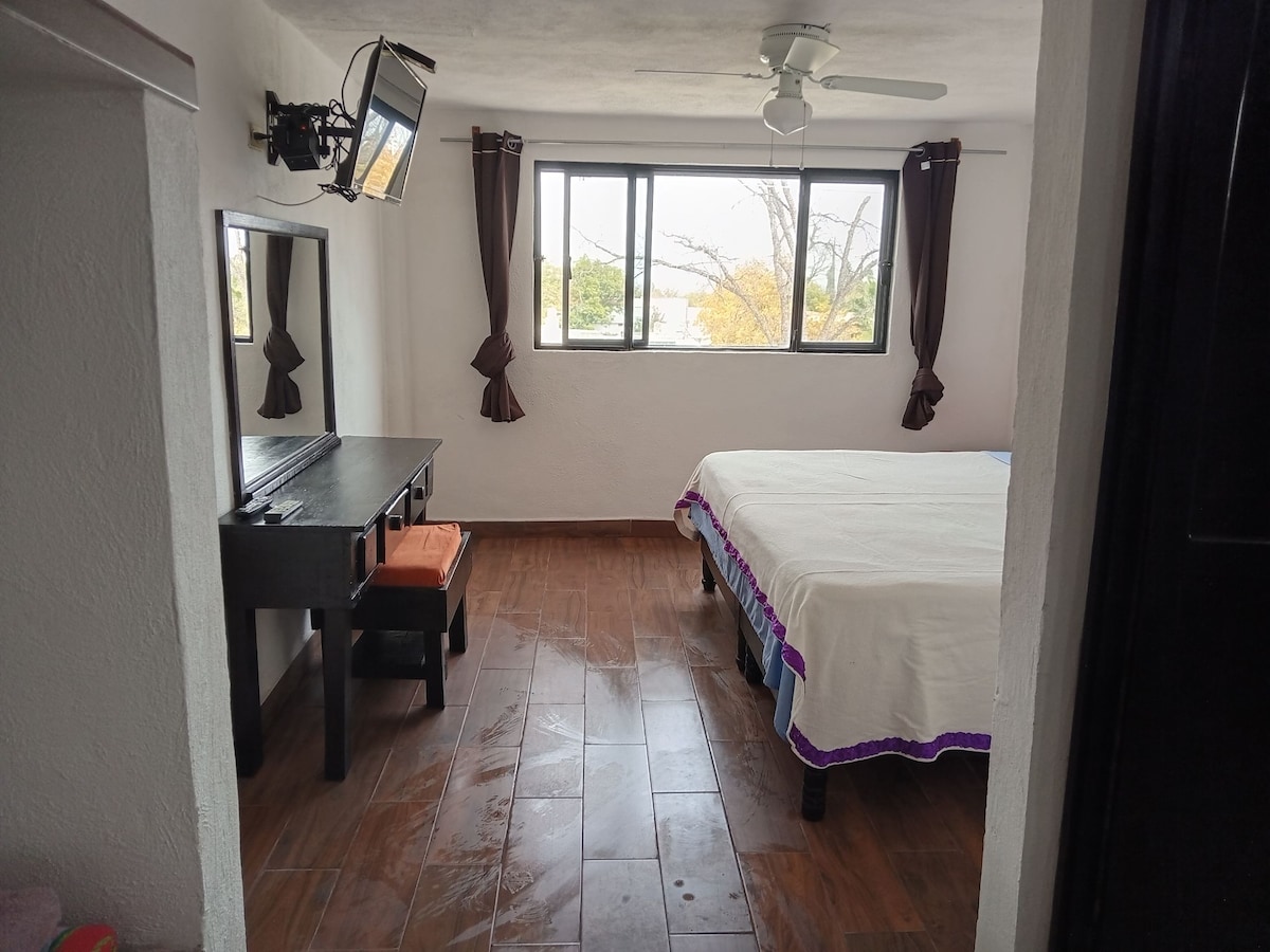 The private room features a spacious bed covered with a white blanket and purple accents. Natural light enters through large windows adorned with brown curtains. A mounted TV is visible on the wall, and a small black table with two stools is positioned nearby.