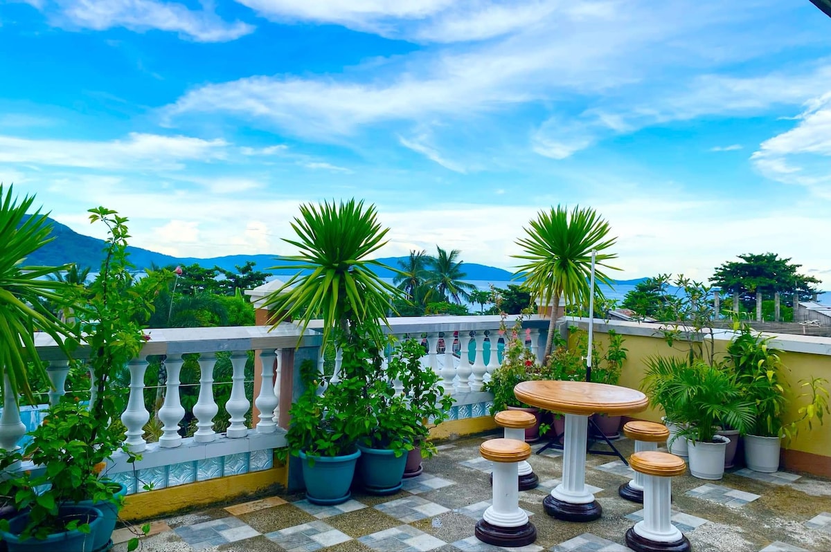 A rooftop patio is adorned with several potted plants and a wooden table surrounded by four stools. The sky showcases soft clouds above distant hills, providing a serene outdoor space ideal for dining or relaxation.