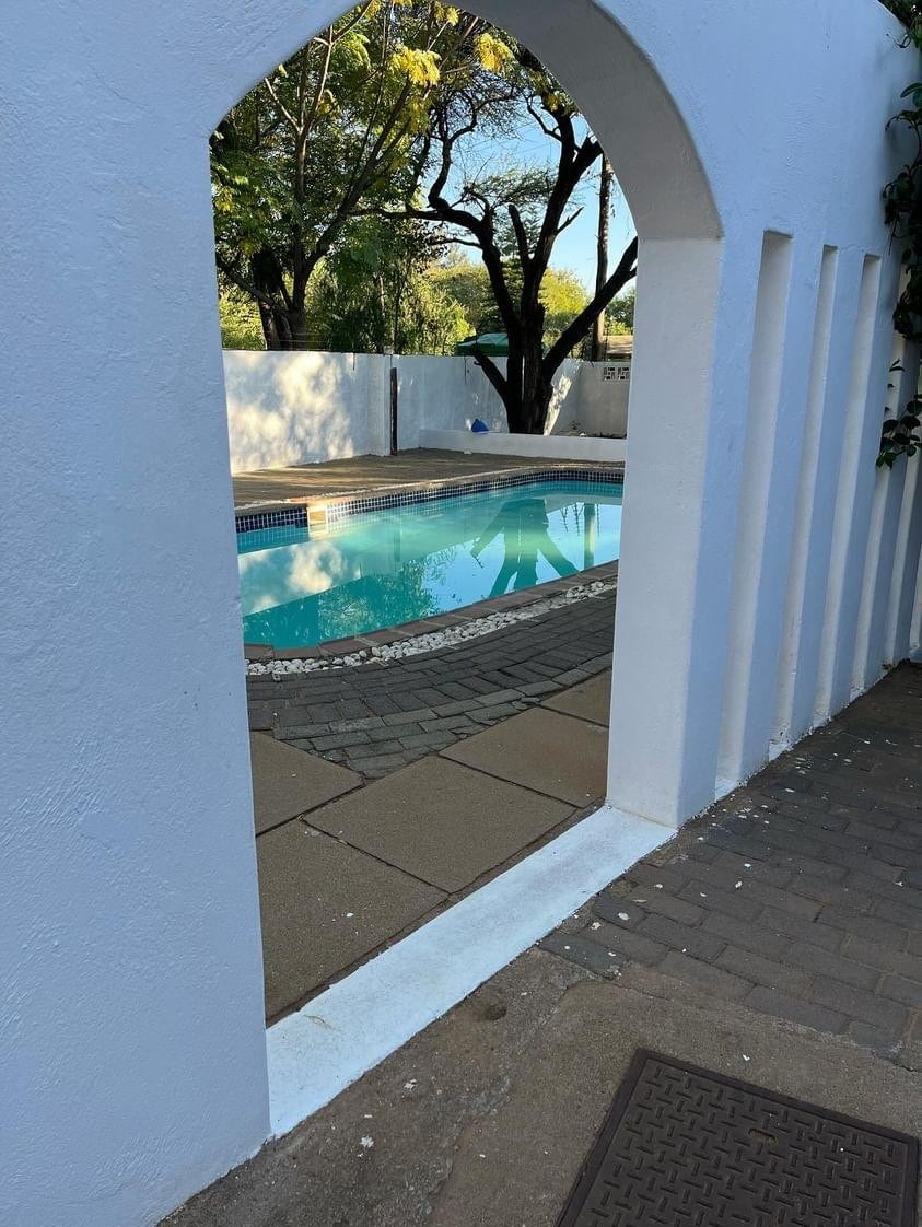 A serene swimming pool is framed by an archway, with sunlight reflecting off the clear water. Surrounding trees provide shade, and a clean, paved area with stones borders the pool. The pool area is enclosed by white walls, contributing to a peaceful setting.