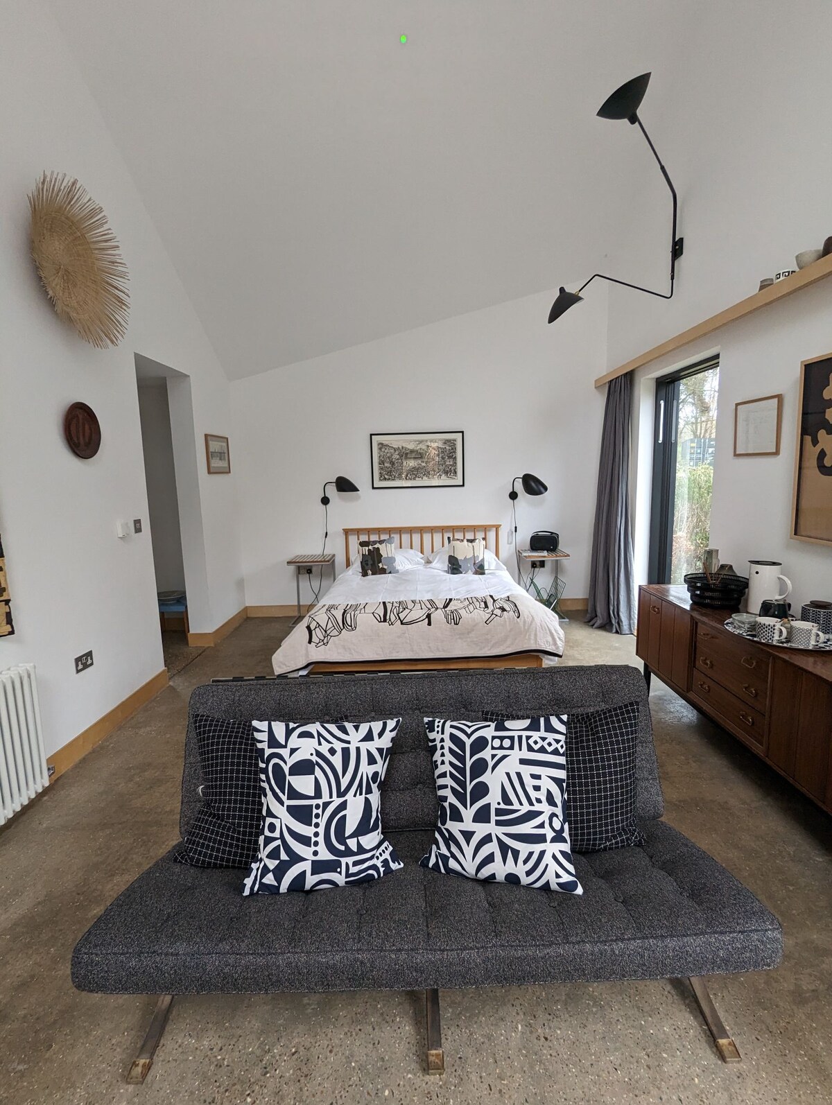 A spacious room features a king-size bed with stylish bedding, positioned beneath a high ceiling. A cozy seating area is furnished with a modern couch adorned with patterned pillows. Soft lighting from the wall-mounted lamps creates an inviting atmosphere, and a sideboard holds various items.
