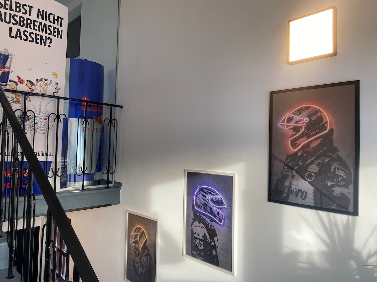 The staircase features racing-themed artwork on the walls, including framed images of a racer in a helmet. A bright square light fixture illuminates the ascent, and the surrounding walls are painted in a neutral tone, enhancing the visual appeal of the artwork.