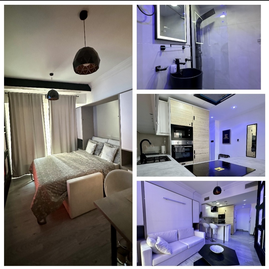 A cozy studio apartment is presented, featuring a spacious bed with soft bedding and an inviting atmosphere. The living area includes a comfortable sofa and a dining table. The kitchen area is fitted with modern appliances, while ambient lighting enhances the overall space.