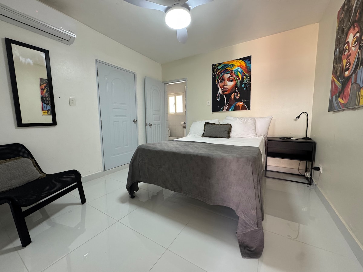 A modern bedroom features a spacious bed dressed in neutral linens, complemented by a bedside table with a lamp. Artistic wall decorations add character. Light-colored walls and tiled floors enhance a clean and airy ambiance, while a ceiling fan provides comfort.