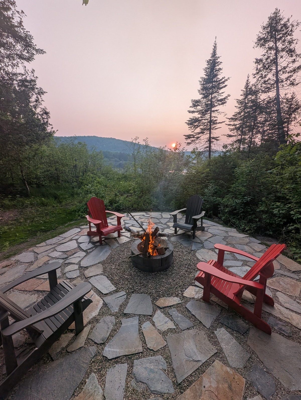 An inviting outdoor fire pit area is surrounded by stone pavers, featuring four colorful adirondack chairs in red and black. Lush greenery frames the scene, while a warm fire is gently flickering in the center. A soft sunset glow is visible in the background.