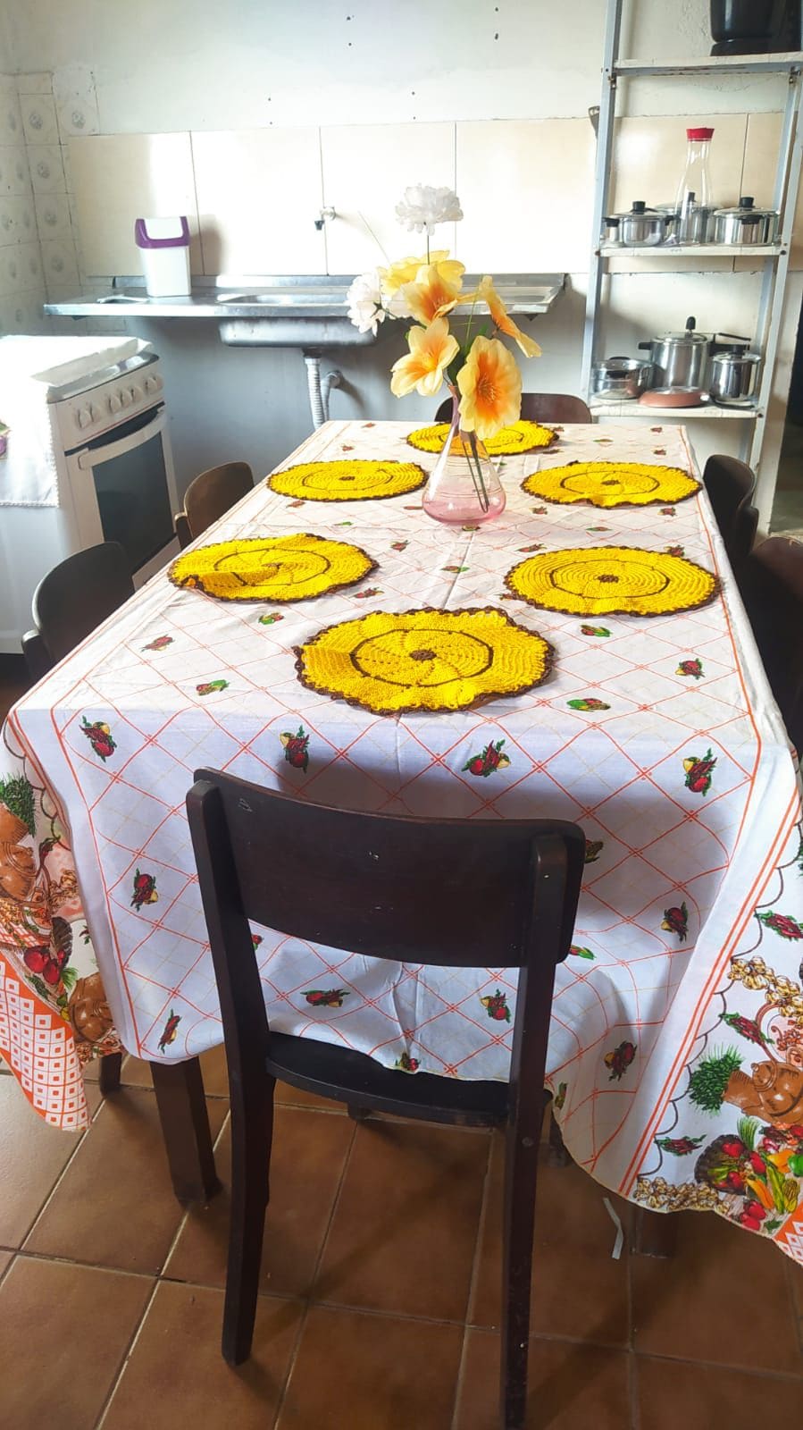 A dining table is set with a white tablecloth featuring floral patterns, adorned with six yellow place mats. A simple vase with orange flowers is positioned at the center. Dark wooden chairs surround the table, and kitchenware is visible in the background.