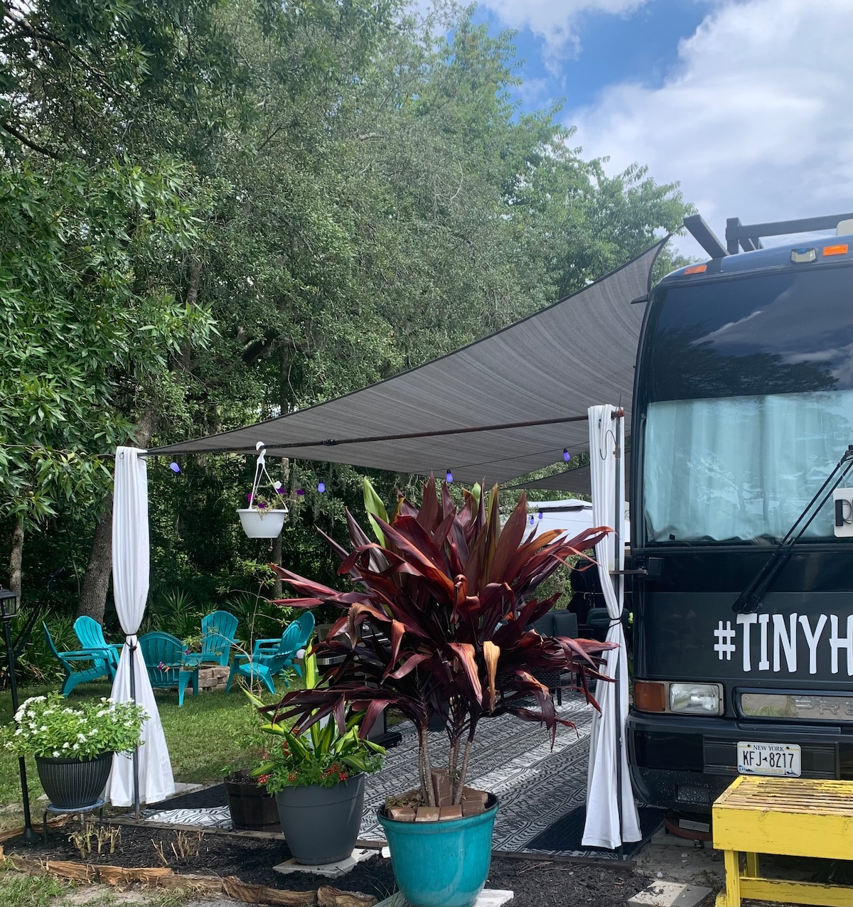 A shaded outdoor seating area features colorful chairs arranged nearby, complemented by vibrant plants. A large awning provides coverage over the space, creating a comfortable atmosphere. A bus adorned with the hashtag #TINYHOUSE is visible, nestled among lush greenery.