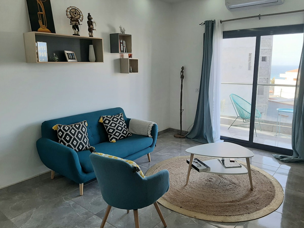 A living area is featured, showcasing a blue sofa with patterned cushions and an area rug beneath a coffee table. A glass door provides access to a balcony, allowing light to fill the space. Decorative shelves are mounted on the wall, enhancing the modern design.