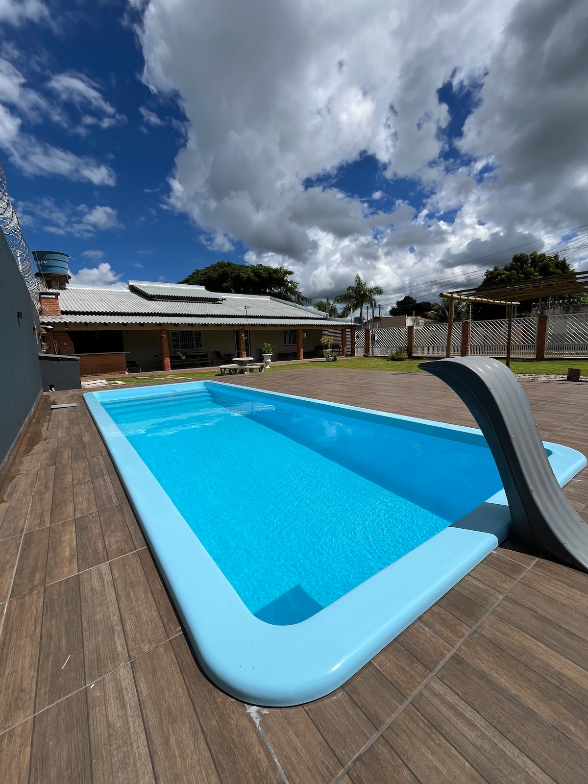 A private rectangular pool is set against a backdrop of a spacious backyard. The pool features clear blue water and a sleek diving board on one side. The surrounding area includes a wooden deck and a grassy lawn, with buildings and trees visible in the distance.