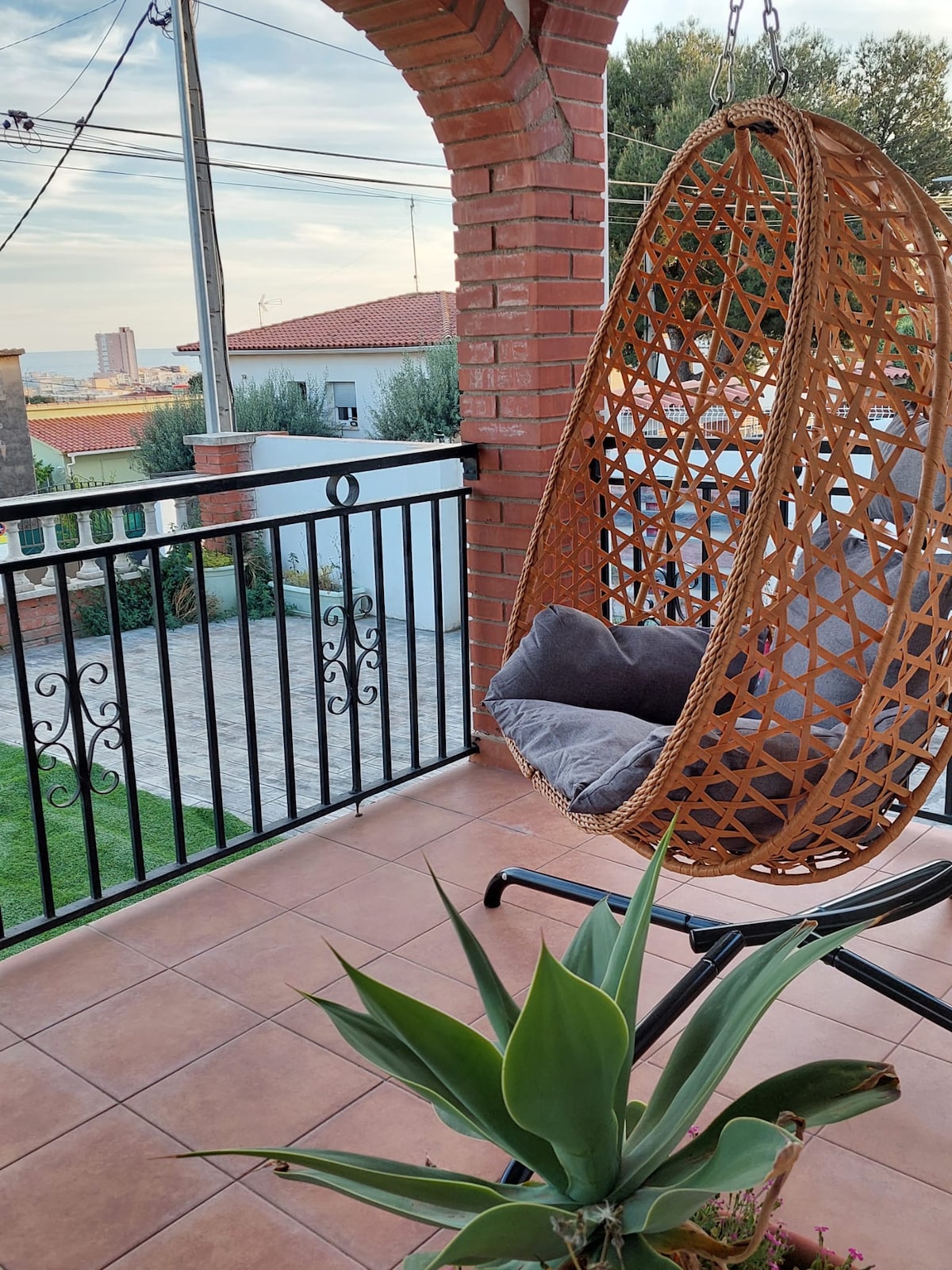A cozy terrace features a woven hanging chair with a cushioned seat. Potted greenery adds a touch of nature, while a view of the neighborhood skyline is visible in the background. The area is framed by decorative wrought iron railings and brick archways.