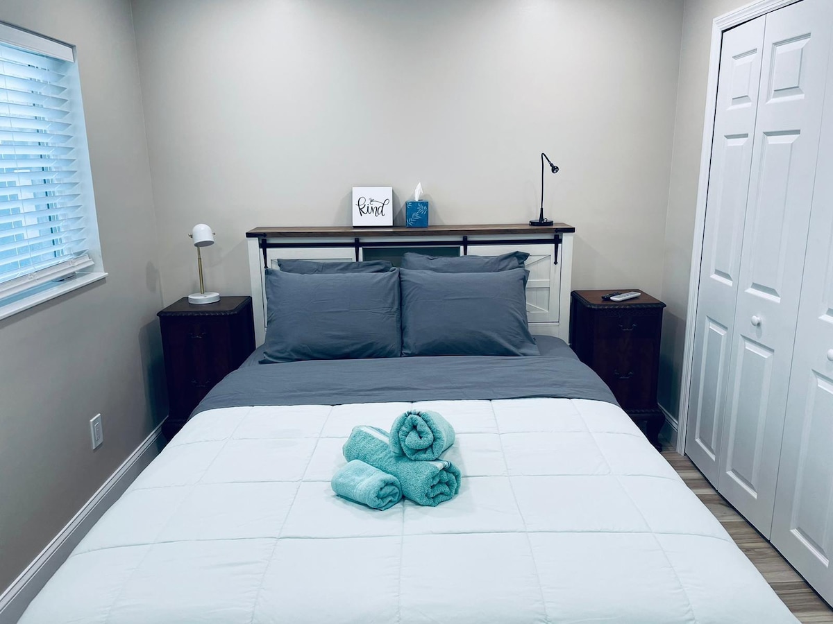A spacious bedroom features a large bed with gray bedding and a white quilted cover. Nightstands are positioned on either side, with a lamp on one and a decorative sign on the headboard. Fresh towels are neatly arranged at the foot of the bed, and a closet is visible.