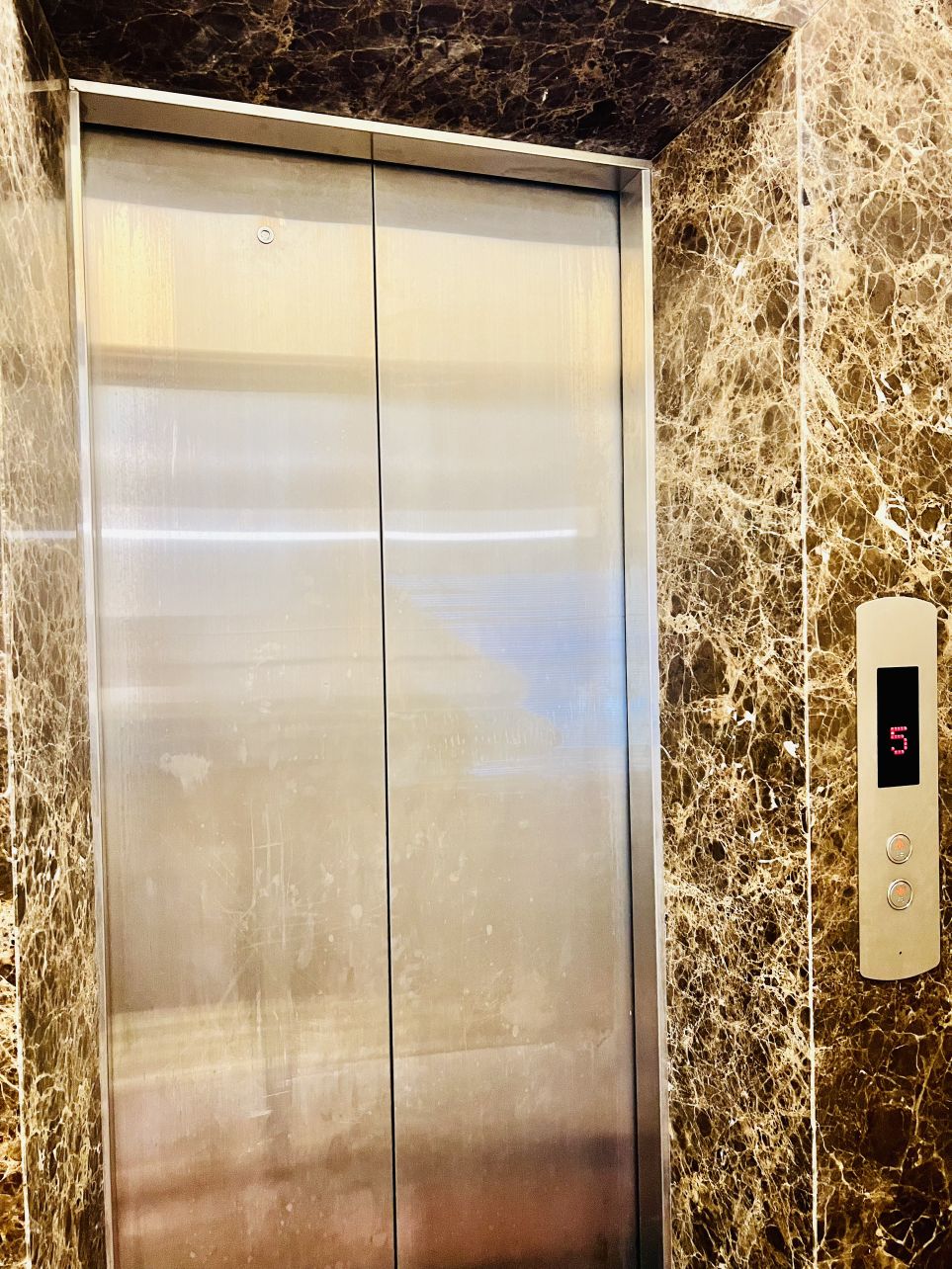 The elevator entrance features a brushed metal door set against a textured, dark marble wall. A digital display and buttons are located beside the door, indicating the current floor and allowing access to various levels.