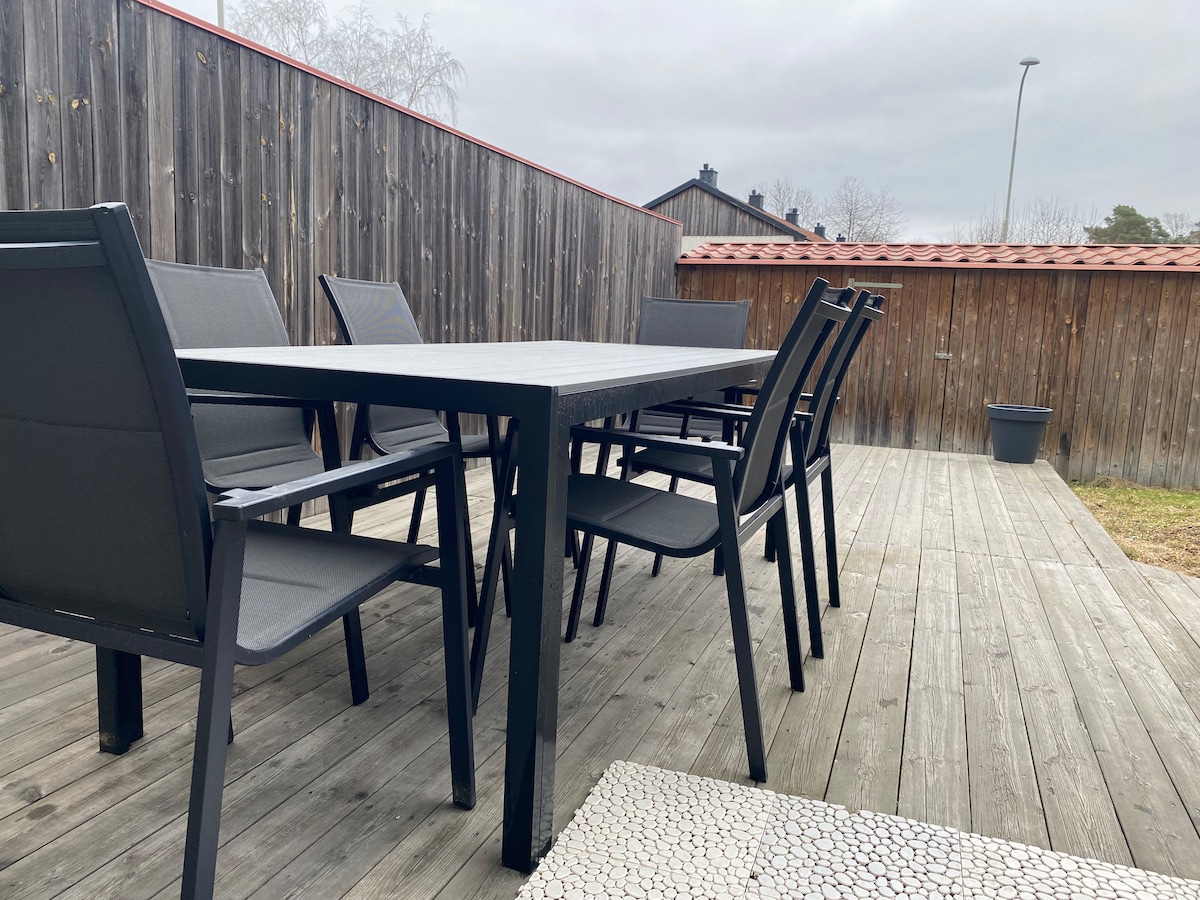 A spacious outdoor dining area features a large dark table surrounded by several gray chairs. The setting is enclosed by wooden fencing, with a wooden deck underfoot and a small potted plant visible in the corner.